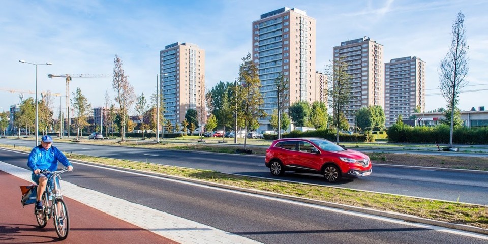 Een straatbeeld met fietser en auto op de voorgrond. In de achtergrond verschijnen grote woonblokken.